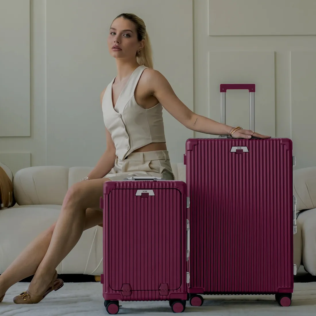 A woman seated beside a burgundy NOBL carry-on and check-in
